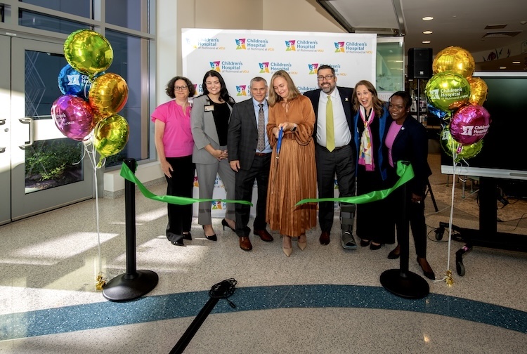 A group dressed in business attire cut a green ribbon inside the new facility. There are colorful balloons around them that say Children’s Hospital of Richmond at VCU.