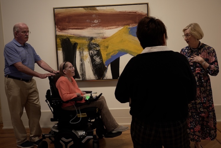 Man pushes his wife in a wheelchair as they listen to tour guides talk about art