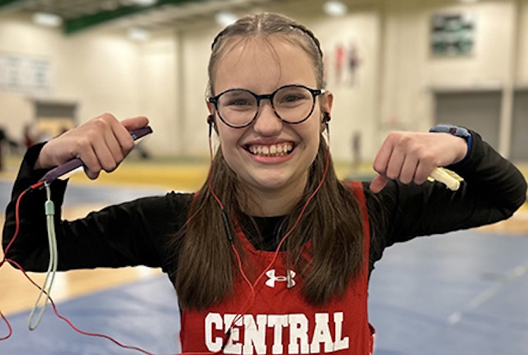 Ali is in a school gym. She is wearing a jersey and holding her arms up to flex her muscles