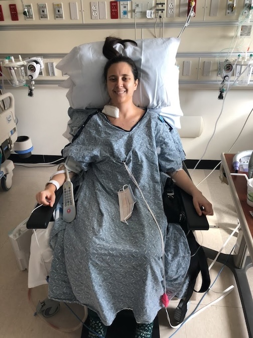 Desiree smiles while sitting in a hospital room and hooked up medical equipment.
