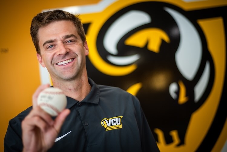 Dr. Brett Goodloe smiles while holding a baseball in front of the VCU Ram logo