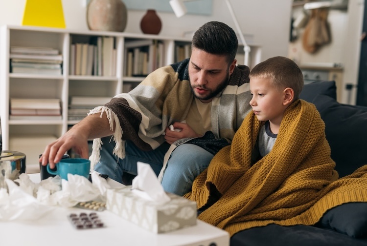 Father and son have the flu. They are at home drinking tea and are covered with blankets.
