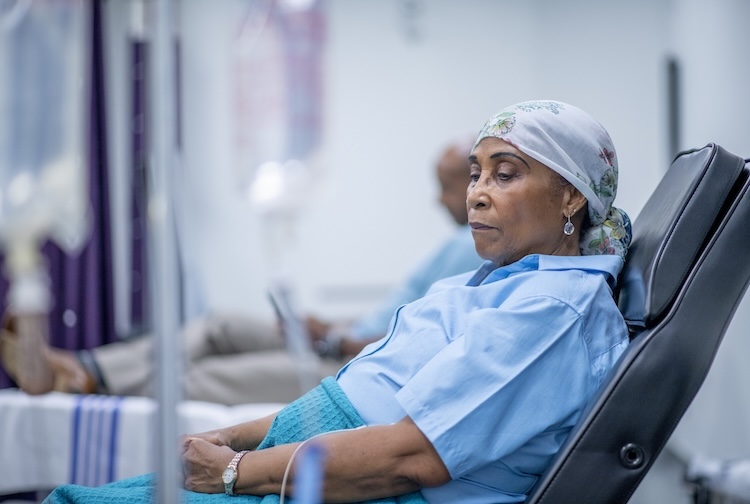 A woman wearing a head sits in a hospital chair with an IV drip next to her.