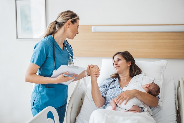 Nurse talks mother about her mental and physical health while holding her hand. The mother is also holding a newborn baby in her other arm.