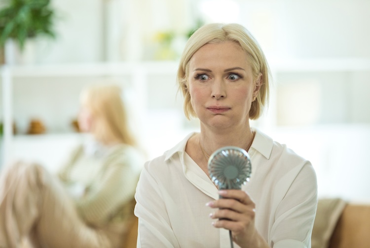 Menopausal woman having a hot flash and using fan. She is displeased her teenage daughter sitting in the background.