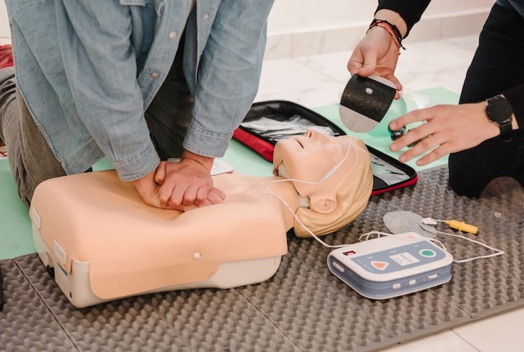 Two people train with a dummy mannequin. One is doing chest compressions for CPR and the other is setting up an AED.