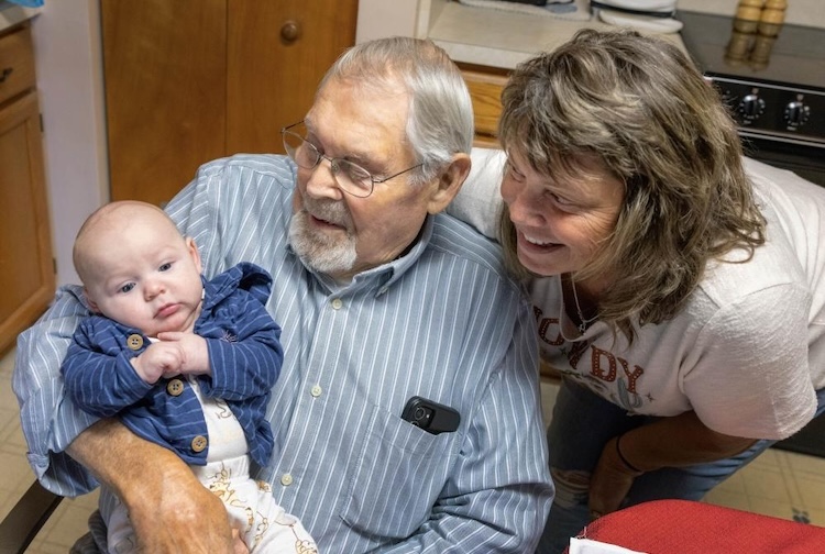 Older man holding baby, and older woman looks on smiling