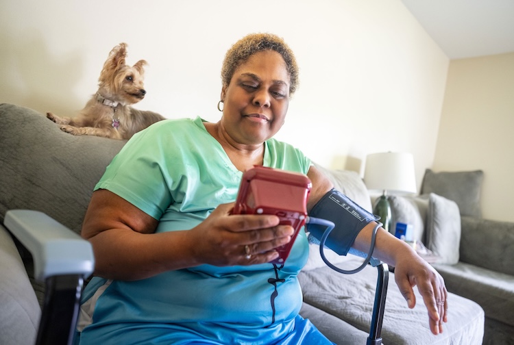 VCU Health patient Ericka looks at a blood pressure cuff for her reading. Her dog sits on the coach looking at her.