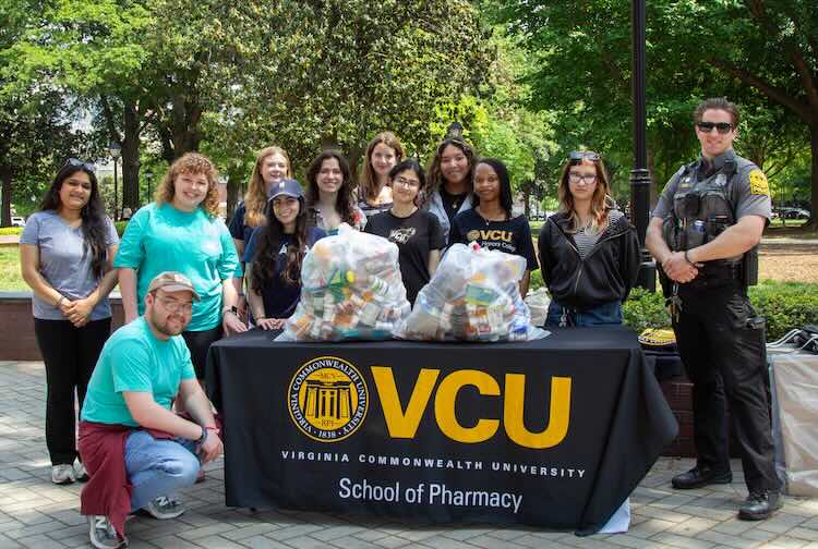 Group of students with a VCU Police Officer standing behind a VCU School of Pharmacy table outside