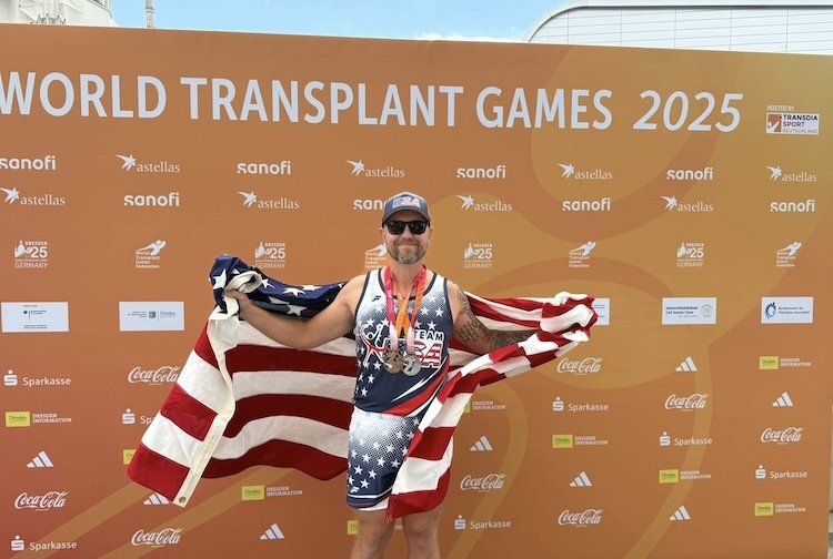 Kyle stands holding a USA flag in front of a sign for the World Transplant Games