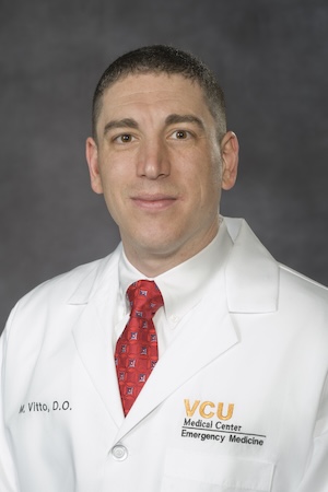 Dr. Vitto wears a white medical coat and a red tie.
