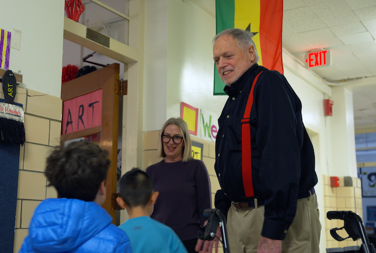 Greg stands next to a teacher and greets students as they walk into the classroom