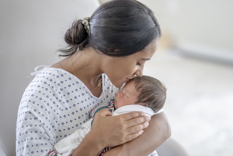 A young mother sits in a rocking chair as she gently kisses her son's forehead. She is wearing a hospital gown and has the baby swaddled in a hospital blanket.