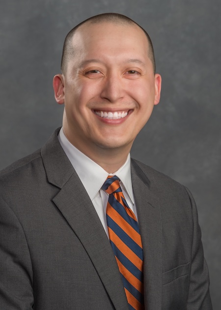 Dr. Melmer wears a white shirt with blue and orange tie along with a grey blazer.