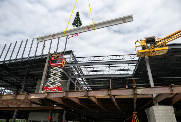 Construction workers hoist a beam up to the top of a structure with an American flag and tree.