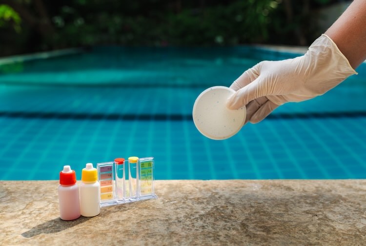 Gloved hand holding chlorine tablet above swimming pool with water testing kit and chemical bottles on stone ledge