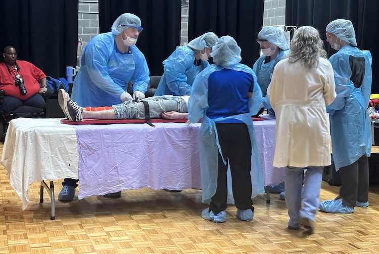 A mock trauma bay set up inside a high school auditorium with medical equipment and volunteers demonstrating emergency care during a Project IMPACT simulation.