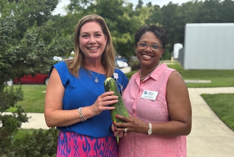Two women pose in front of a garden holding a fresh zucchini.
