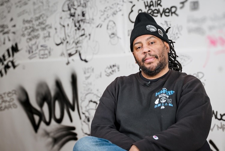 Artist sits in front of a wall filled with graffiti.