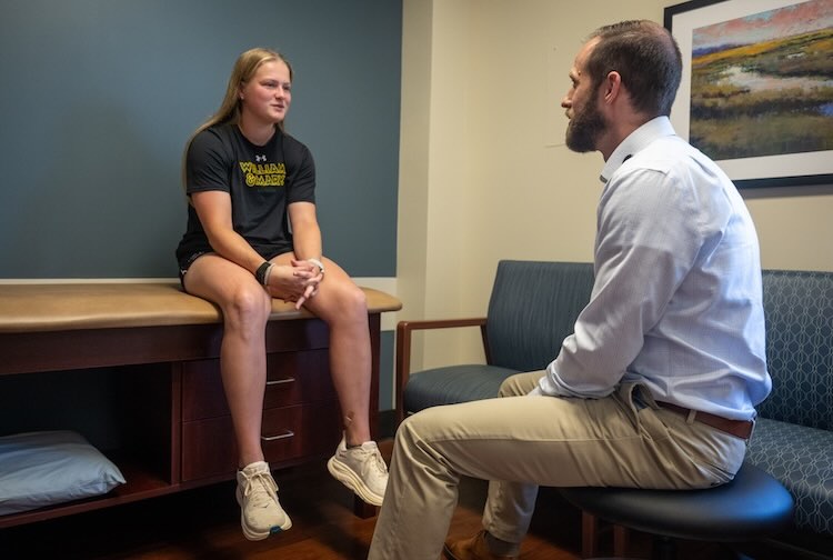 Lacrosse player sits on patient exam bed and talks to sports medicine doctor