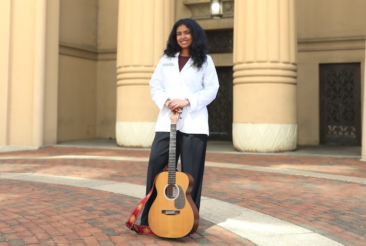 Sravani standing outside of building in her white coat with guitar in hand.