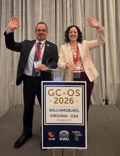 Two VCU Health team members in suits wave their hands and smile behind a sign for the summit.