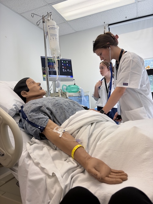 High schoolers take a blood pressure reading with a manikin laying in a hospital bed.