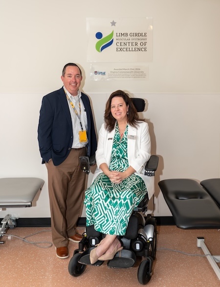 Kathryn wears a green and blue dress while sitting in a wheelchair next to Dr. Johnson, who is in a suit. They are smiling next to the plaque for the LGMD Center of Excellence.