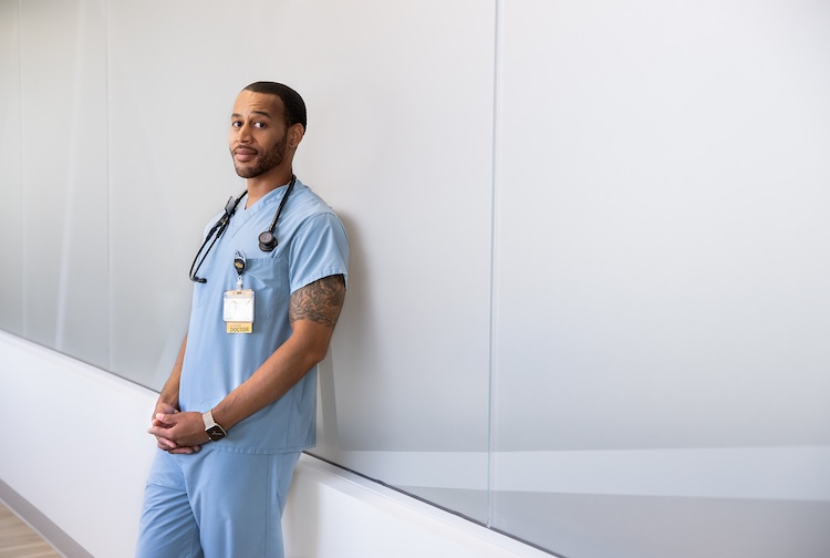 Doctor in scrubs leans up against a wall.