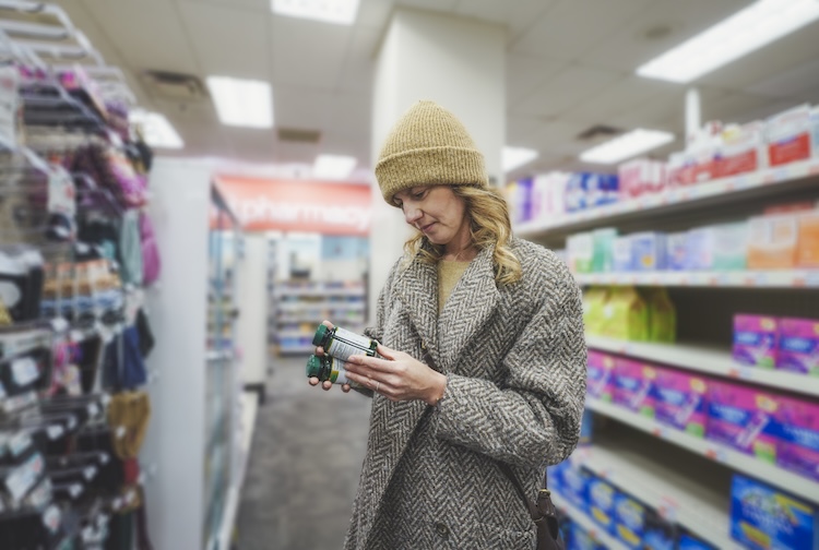 A woman, wearing a winter coat and hat, is shopping for multivitamins in an aisle of a pharmacy