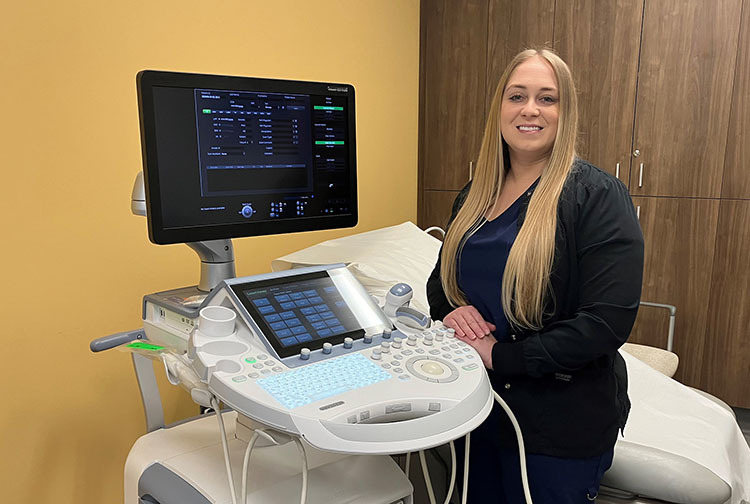 A lady wearing a black medical uniform poses next to an ultrasound machine.