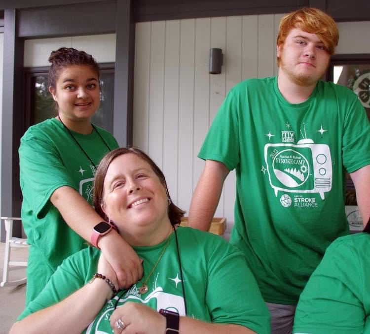 Mom sitting down holding her daughter's hand. Son standing next to them smiling. They are all wearing green shirts.
