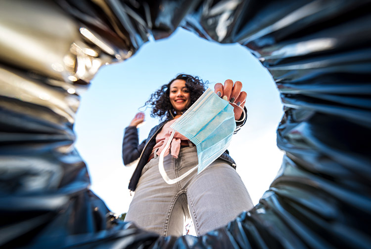 a smiling lady drops a mask in a trash can.
