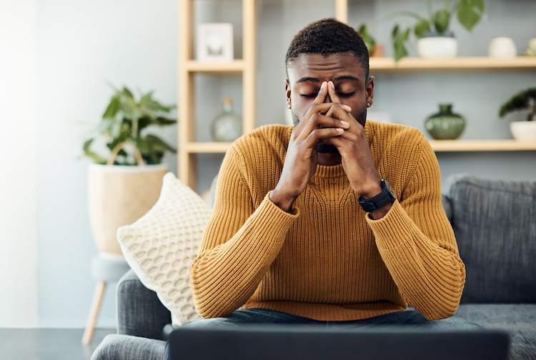 Image of a young man looking stressed out at home. His hands are infront of his face while he sits on a couch.