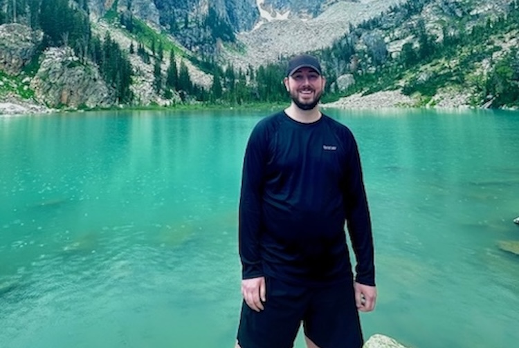 Joey stands in front of a large mountain and lake.