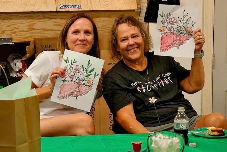 two women hold up colored in drawings of livers with flowers growing out of them