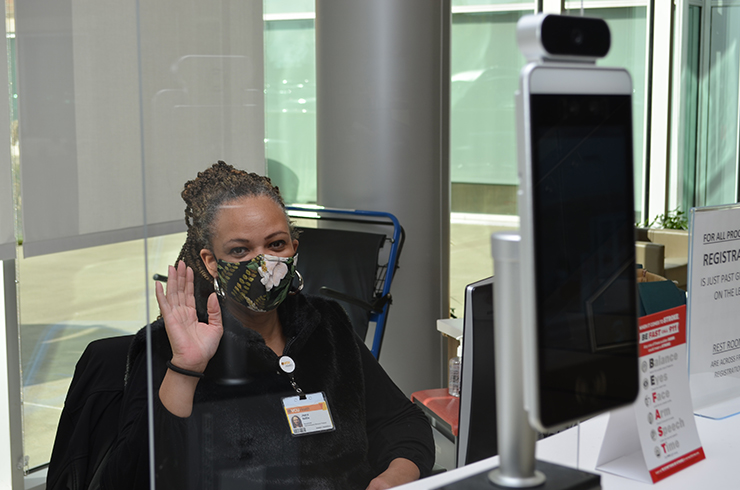 Joyce Nolli waves from behind the divider wall at a temperature screening station at VCU Health CMH.