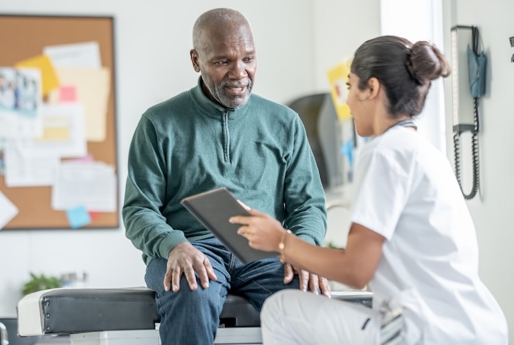 A senior African American man sits on an examination table as a nurse reviews information on a tablet in a bright clinic
