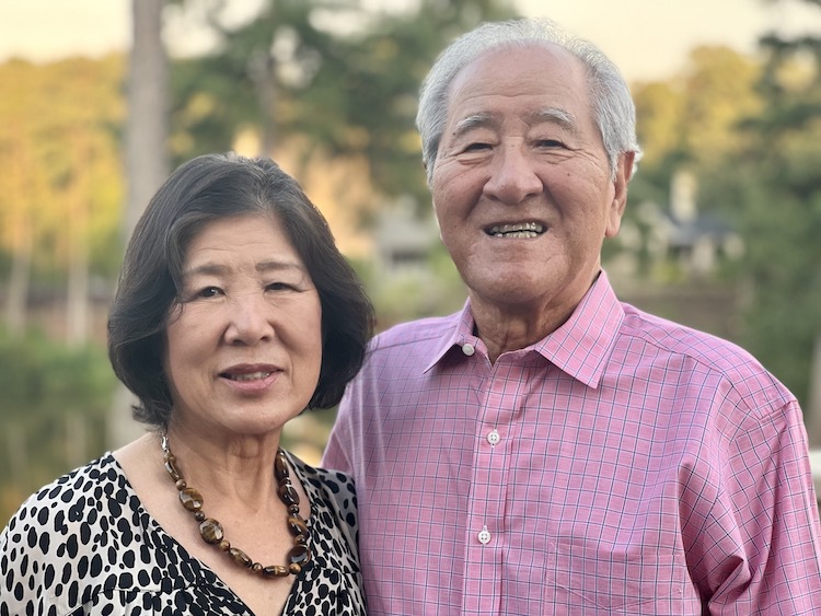 Two older adults stand next to each other smiling.