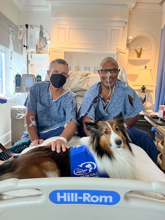 Two men standing sitting in hospital gowns next to one of the VCU Dogs on Call therapy dogs.