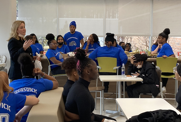 lady with blonde hair wearing black blazer speaks to students who are all wearing blue shirts. 