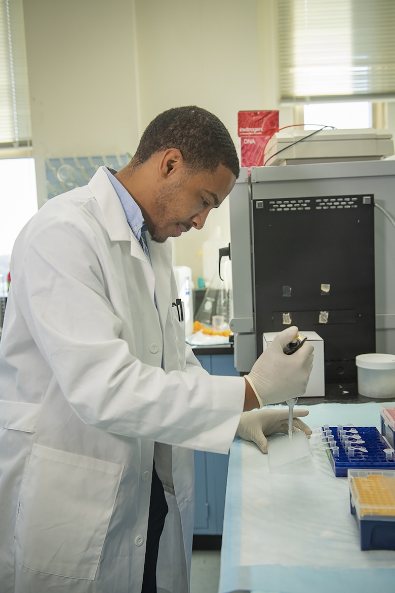 Man in white lab coat is using a pipette to drop a liquid into a tube.
