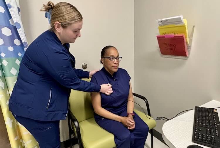 Victoria connects a microphone to a patient to prepare them for a virtual appointment in a hospital room.