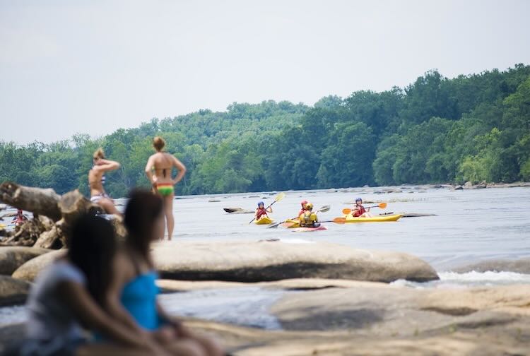 People in bathing suits sitting and standing on rocks along the river as kayakers go by.