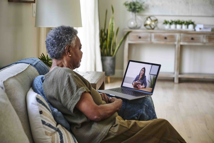 A woman sits on a couch and looks at her laptop screen while having a video call with a health care provider.