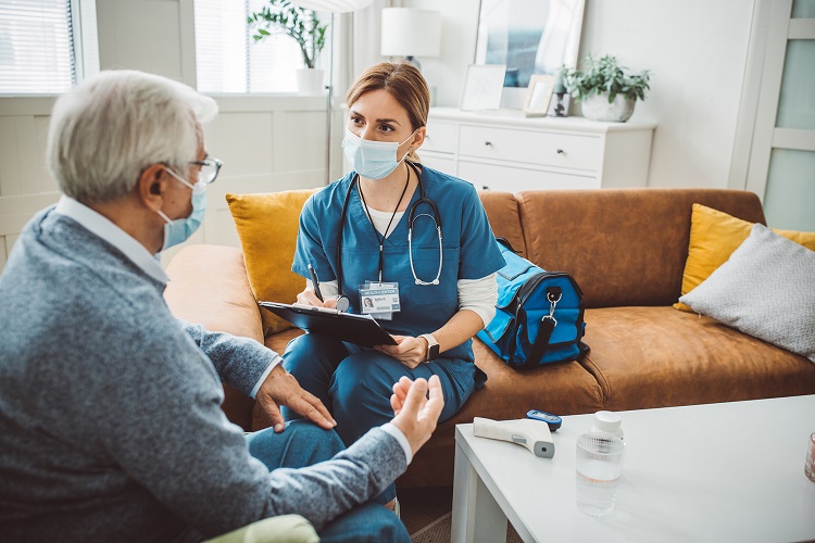 Woman in scrubs and mask sitting on a couch writing on a clipboard while talking to elderly man wearing a mask