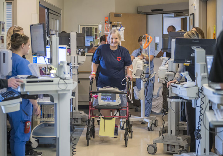 A woman walks down a hospital hallway using a walker and attached to medical equipment. 